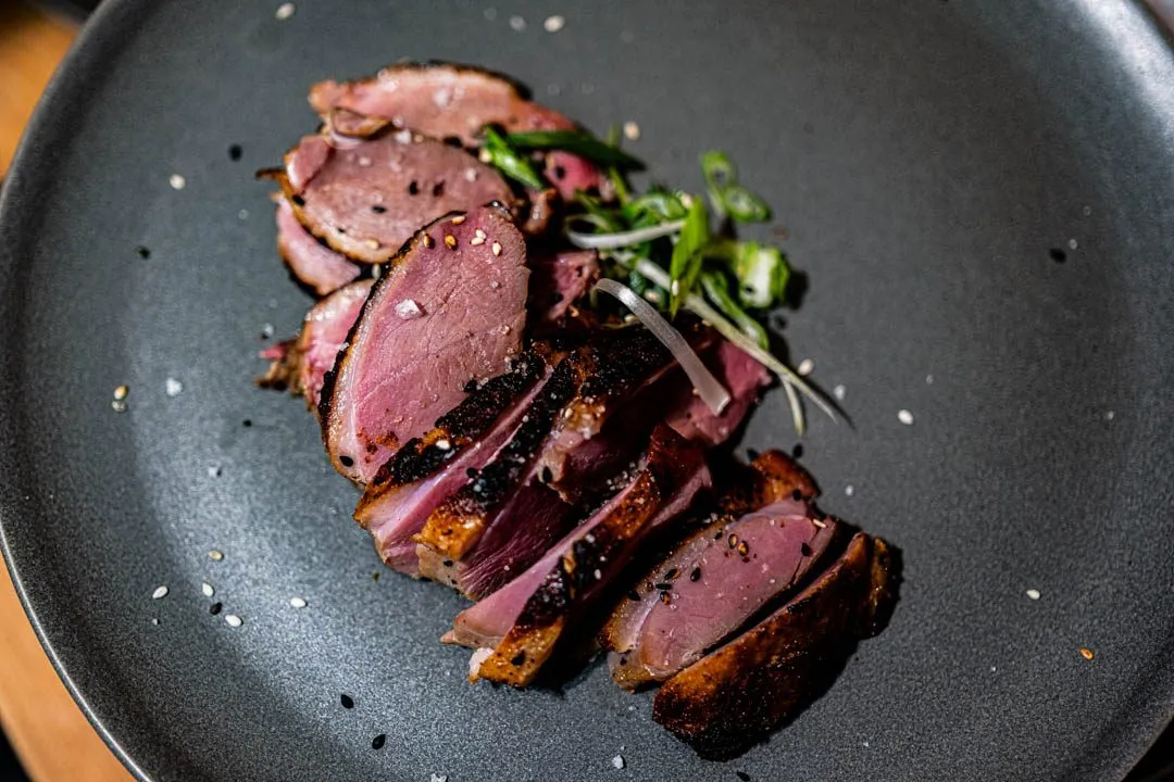 Sliced miso-cured duck breast with shatter-crisp golden skin fanned on a plate, topped with julienned green onions and flaky salt