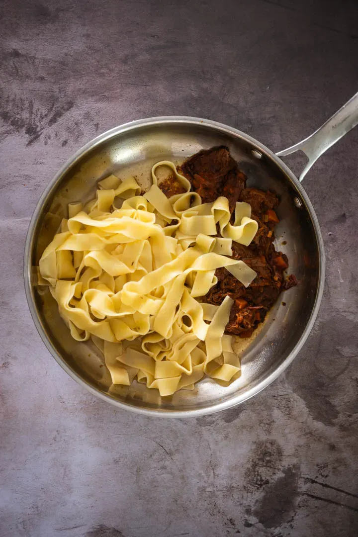 Wide pappardelle ribbons being tossed with the slow-cooked beef ragu in a pan