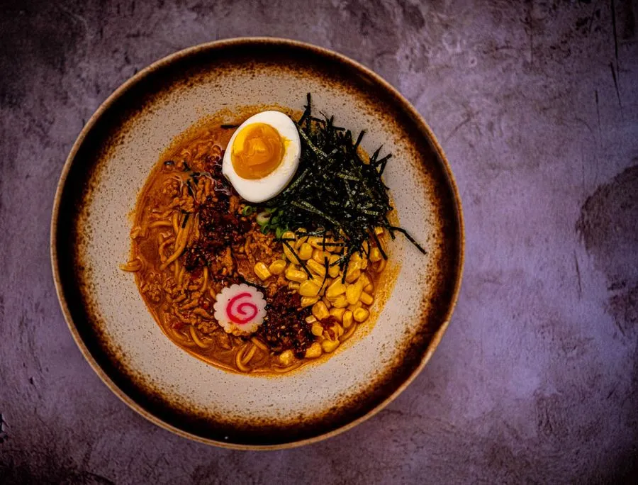 Close-up of a bowl of spicy miso ramen with scallions, nori sheets, and a drizzle of vivid garlic-togarashi oil pooling on top of the broth