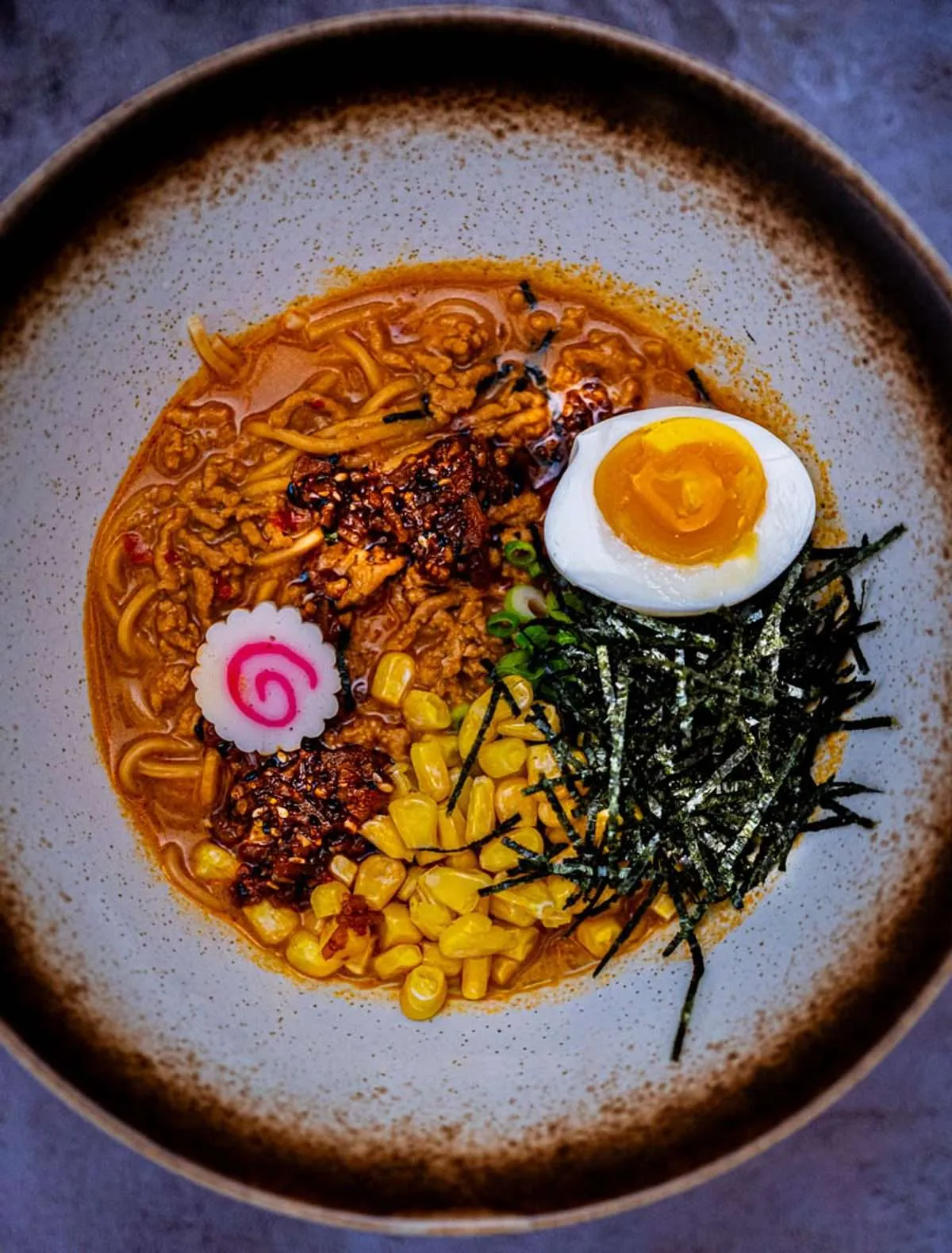 Two bowls of bold spicy miso ramen with ground pork, garlic-togarashi oil, scallions, and nori on a dark wood table