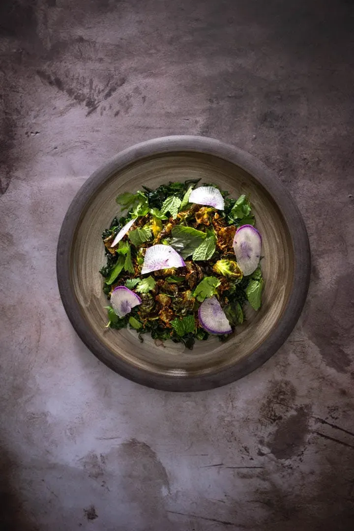 Shatter-crisp fried Brussels sprout leaves piled over dark shaved kale, bright herbs, and paper-thin purple daikon on a white plate