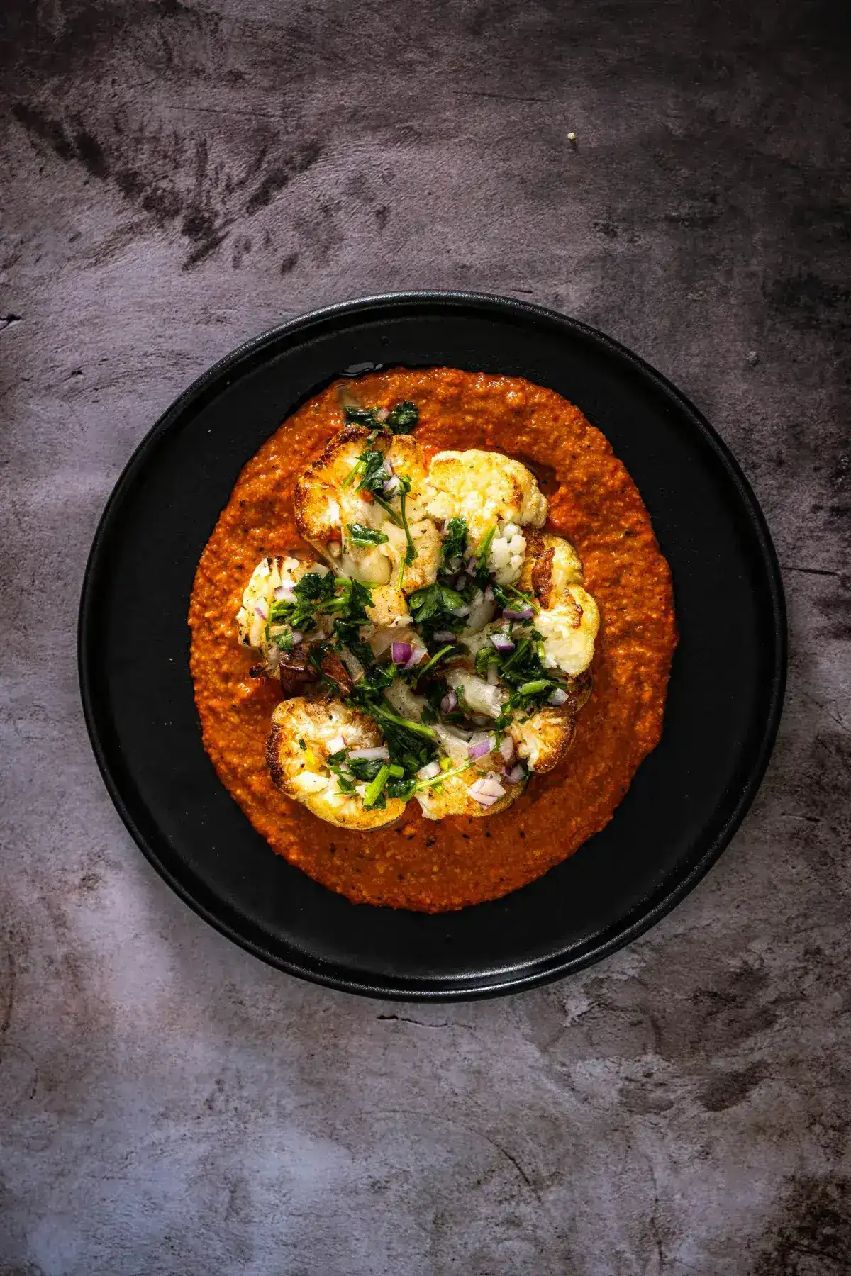 Golden roasted cauliflower steaks on a plate drizzled with smoky red romesco sauce and fresh parsley dressing