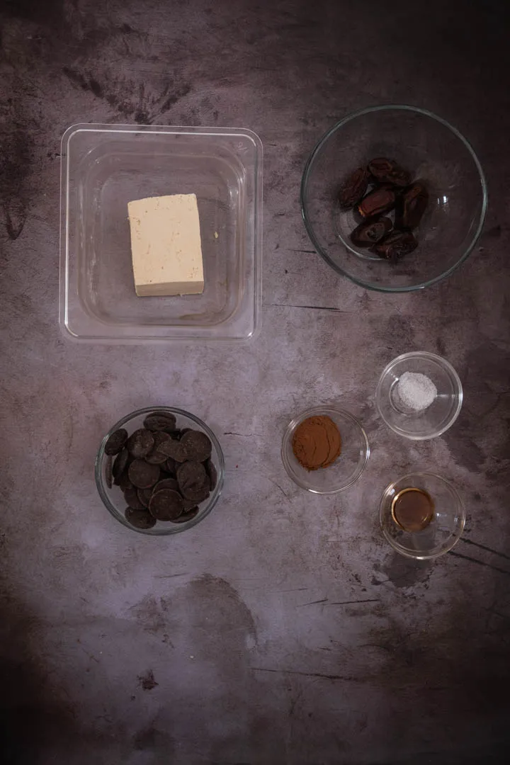 Ingredients for chocolate tofu pudding laid out: silken tofu, dates, milk chocolate, cacao powder, olive oil, and vanilla