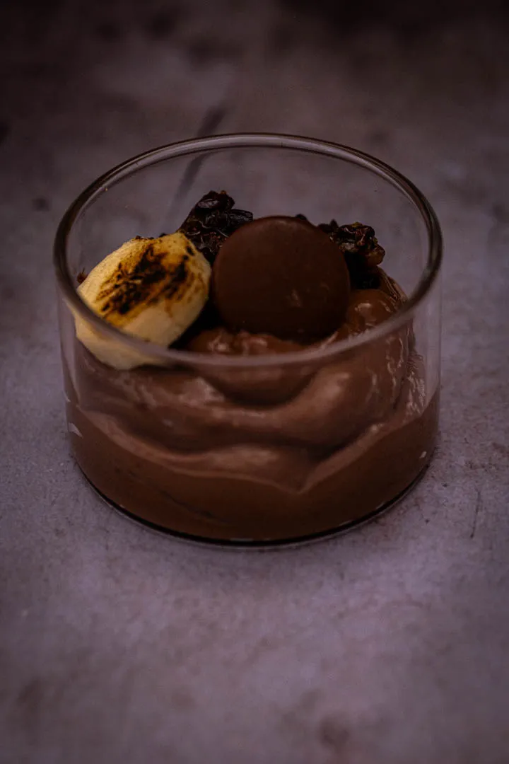 Close-up of finished chocolate tofu pudding with flaky salt, candied cacao nibs, and banana on a dark surface
