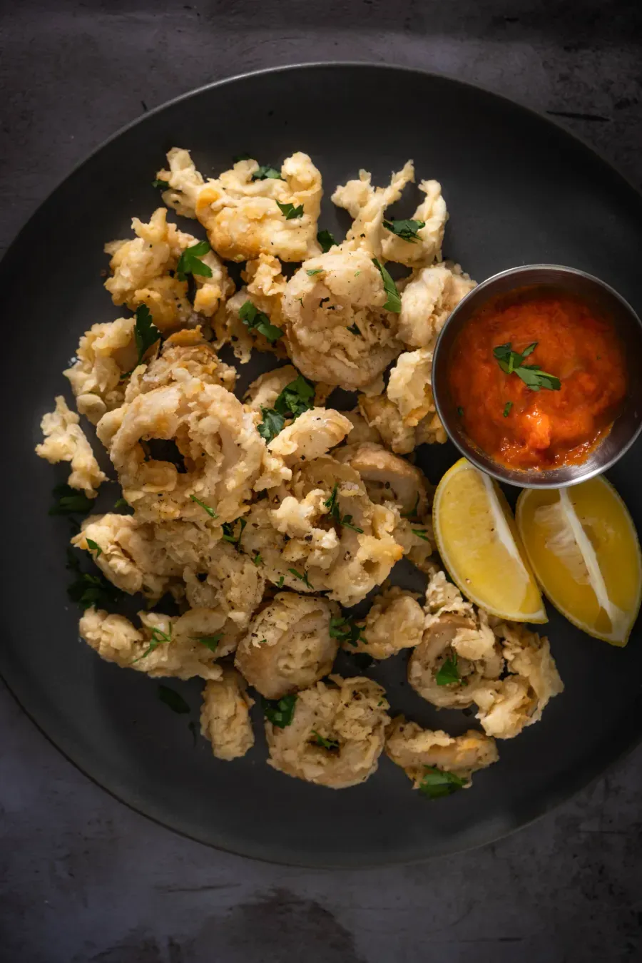 Tumble of golden-fried vegan calamari rings glistening with oil, stacked alongside bright lemon wedges on a warm plate