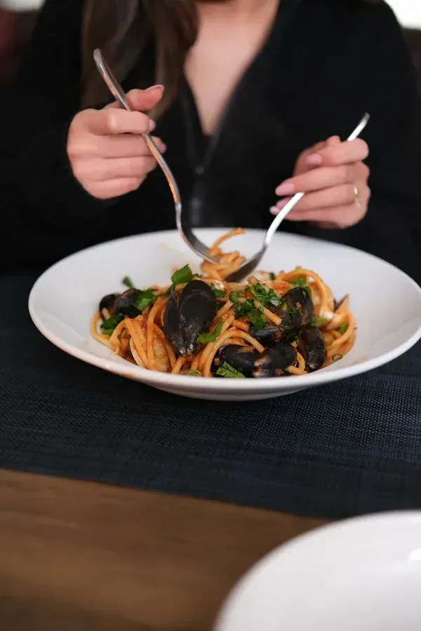 Warm overhead scene of someone twirling spicy bucatini from a wide bowl, mussels and herbs scattered around the rim, candlelight catching the glossy sauce