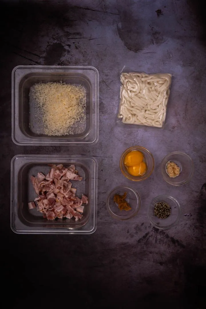 Ingredients for miso udon carbonara laid out on a dark surface: grated Parmesan, frozen udon, diced bacon, egg yolks, miso paste, and black pepper