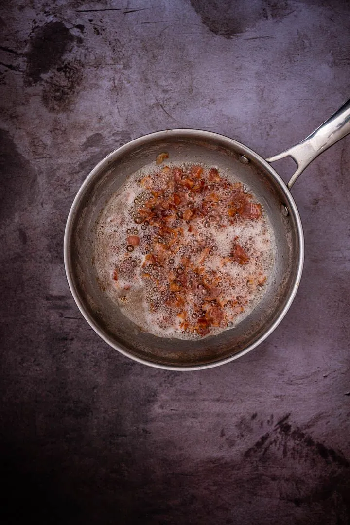 Diced bacon rendering in a stainless steel pan, fat bubbling and pieces turning golden and crisp