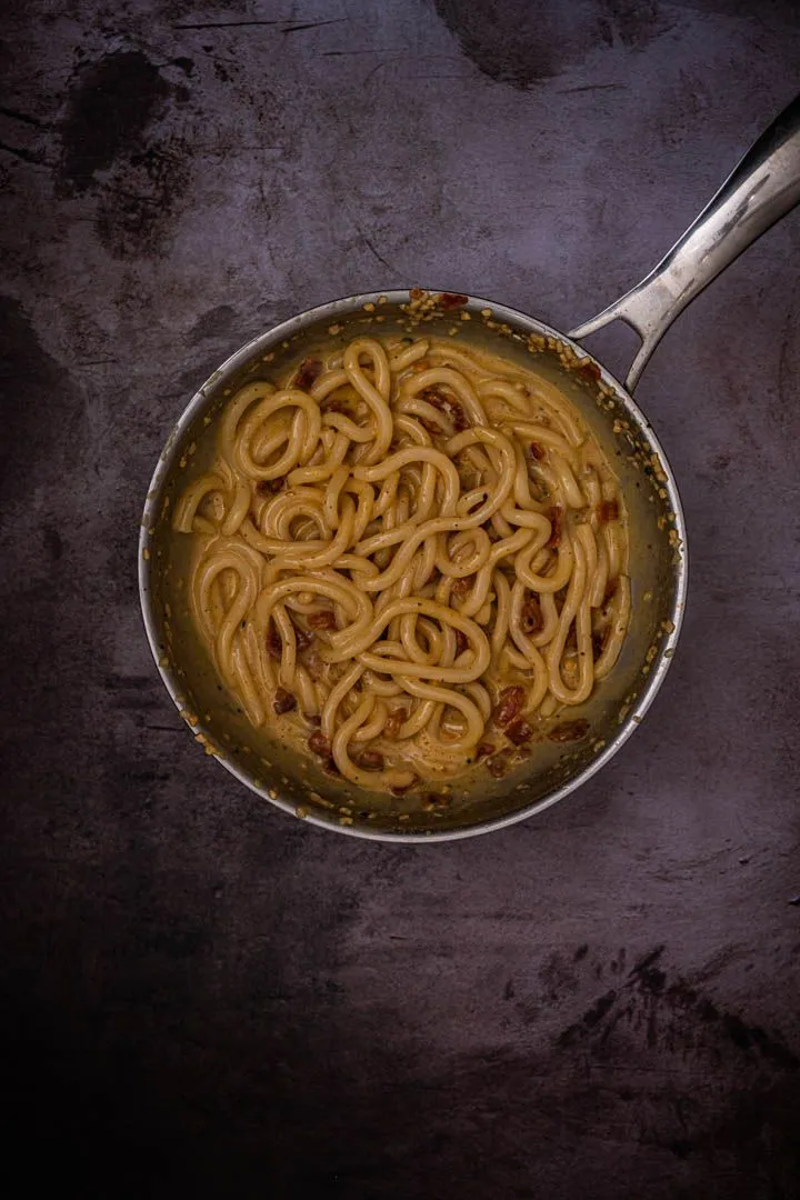Glossy udon noodles fully coated in the silky carbonara sauce in a stainless steel pan, the emulsion shiny and clinging to every strand