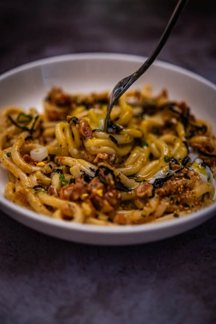 Close-up of finished miso udon carbonara in a white bowl with a fork lifting noodles, crispy bacon, chives, and nori visible throughout
