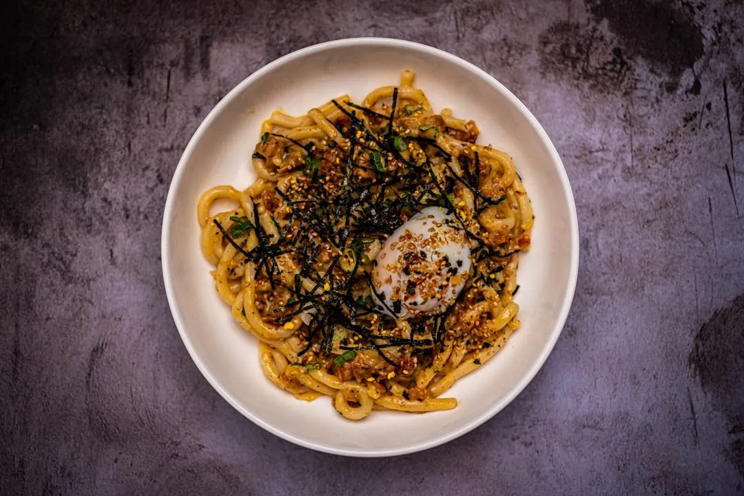 Overhead shot of miso udon carbonara in a white bowl topped with nori, furikake, and a soft egg on a dark surface