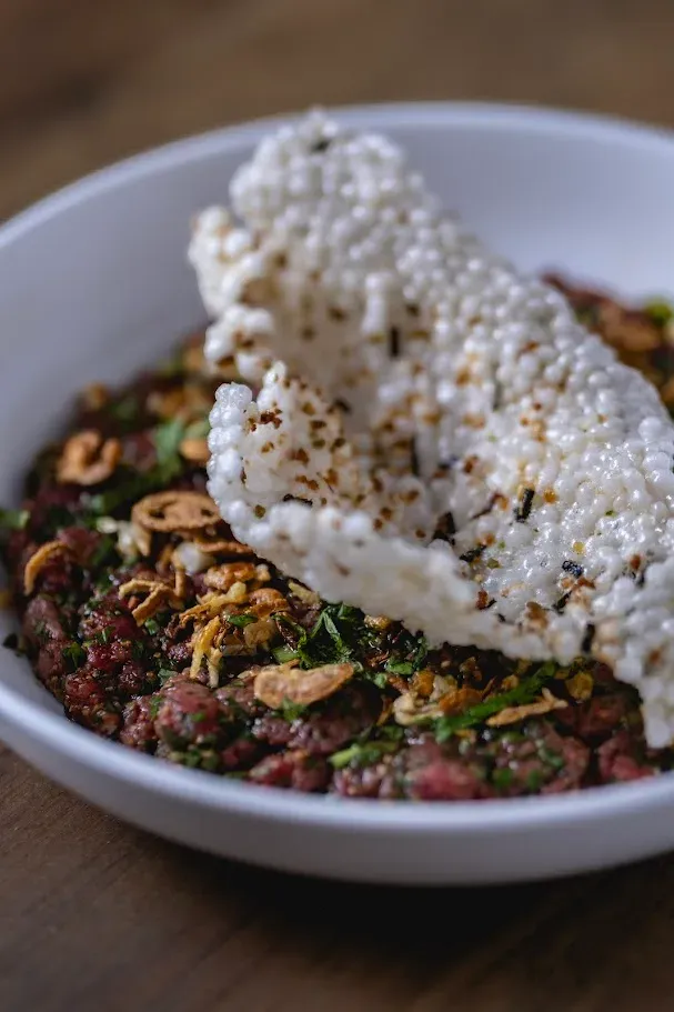 Northern Thai beef tartare in a wide bowl, covered in fresh herbs, crispy shallots, and garlic chips, surrounded by Boston lettuce cups