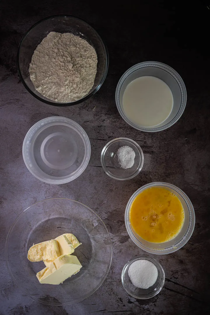 Choux pastry ingredients measured out: water, milk, butter pieces, sugar, salt, bread flour, and beaten eggs in separate bowls
