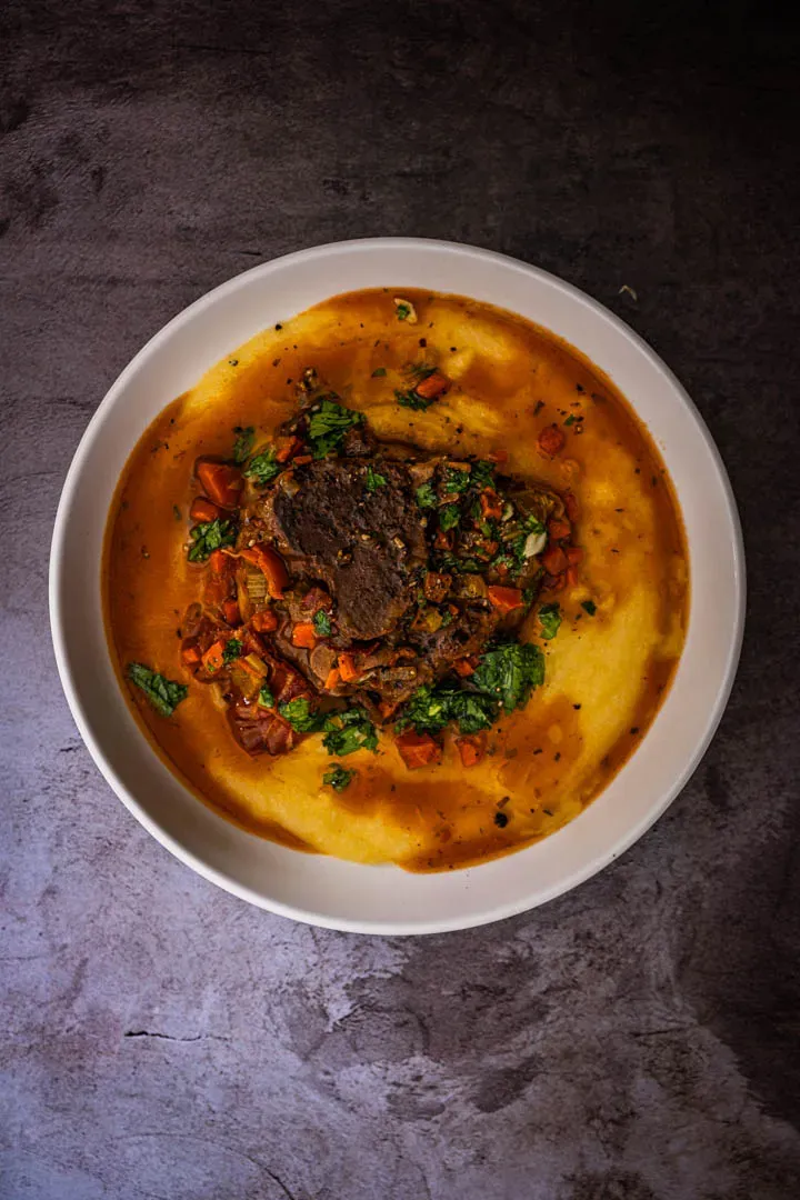 Pork osso buco plated on creamy polenta with braising sauce ladled over the shank and bright green gremolata on top