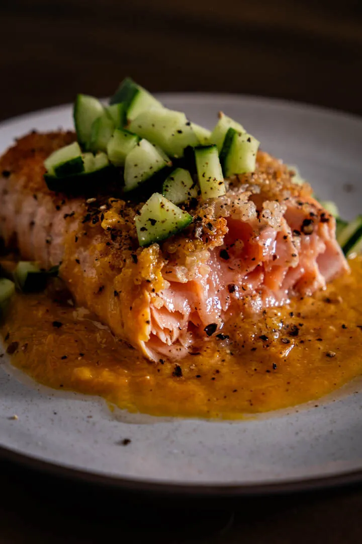 Quinoa-crusted salmon fillet drizzled with spicy orange miso sauce and scattered with fresh diced cucumber on a white plate