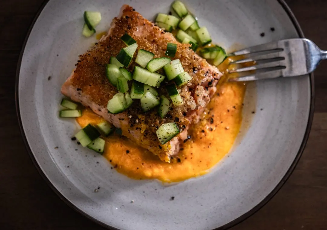 Golden quinoa-crusted salmon fillet glistening with spicy orange miso sauce, scattered with cool diced cucumber on a sleek white plate