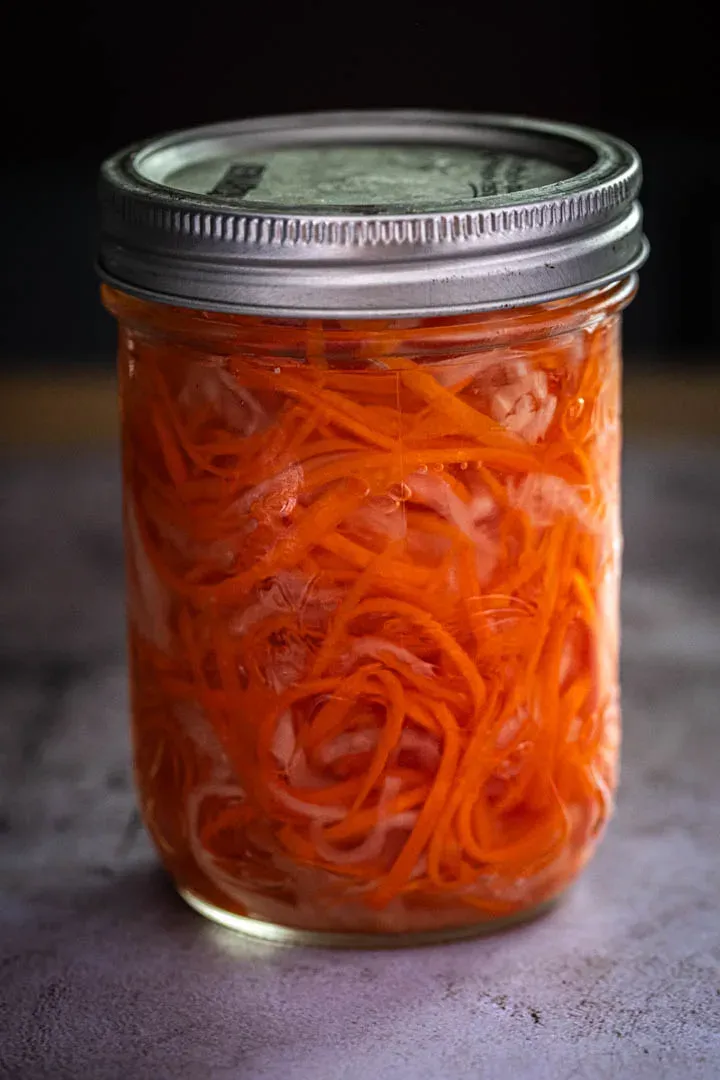 Glass jar packed with Vietnamese do chua, julienned daikon and carrot matchsticks glowing orange and white in clear sweet-tangy brine